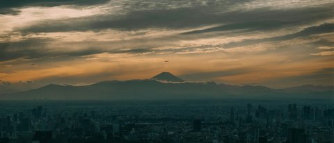 Mount Fuji's dark silhouette floats above Tokyo's endless grid of towers at dusk, the sky melting from peach to indigo as the city's lights begin to flicker on