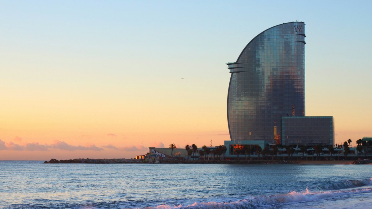 The sail-shaped W Barcelona rising over Barceloneta beach at dusk, pastel peach sky melting into a Mediterranean horizon as surf curls onto the darkening sand