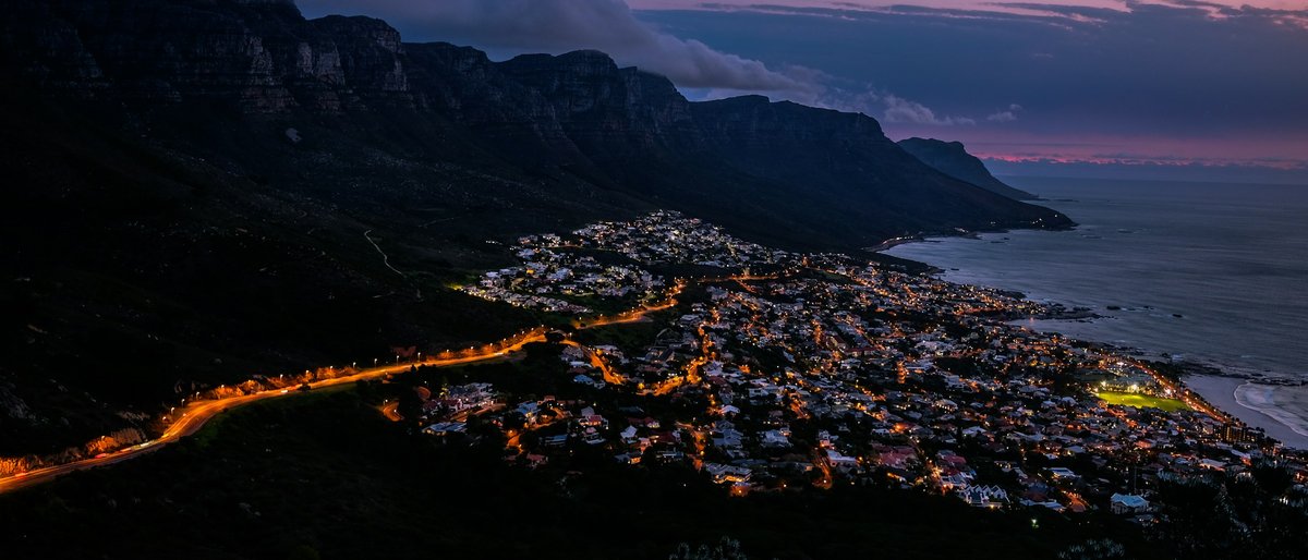 Camps Bay glows below the Twelve Apostles ridge at violet twilight, warm street-lamp ribbons threading dark coastal suburbs while low cloud spills over the cliffs against pink-mauve sky