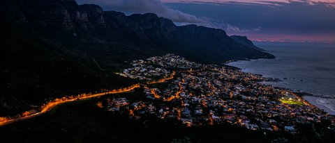 Camps Bay glows below the Twelve Apostles ridge at violet twilight, warm street-lamp ribbons threading dark coastal suburbs while low cloud spills over the cliffs against pink-mauve sky