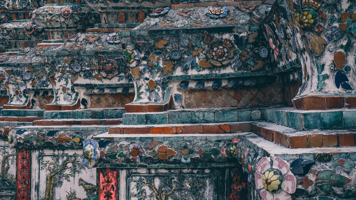A weathered close-up of Wat Arun's porcelain mosaic, broken china flowers and faded blue glaze stacked into the temple's flank