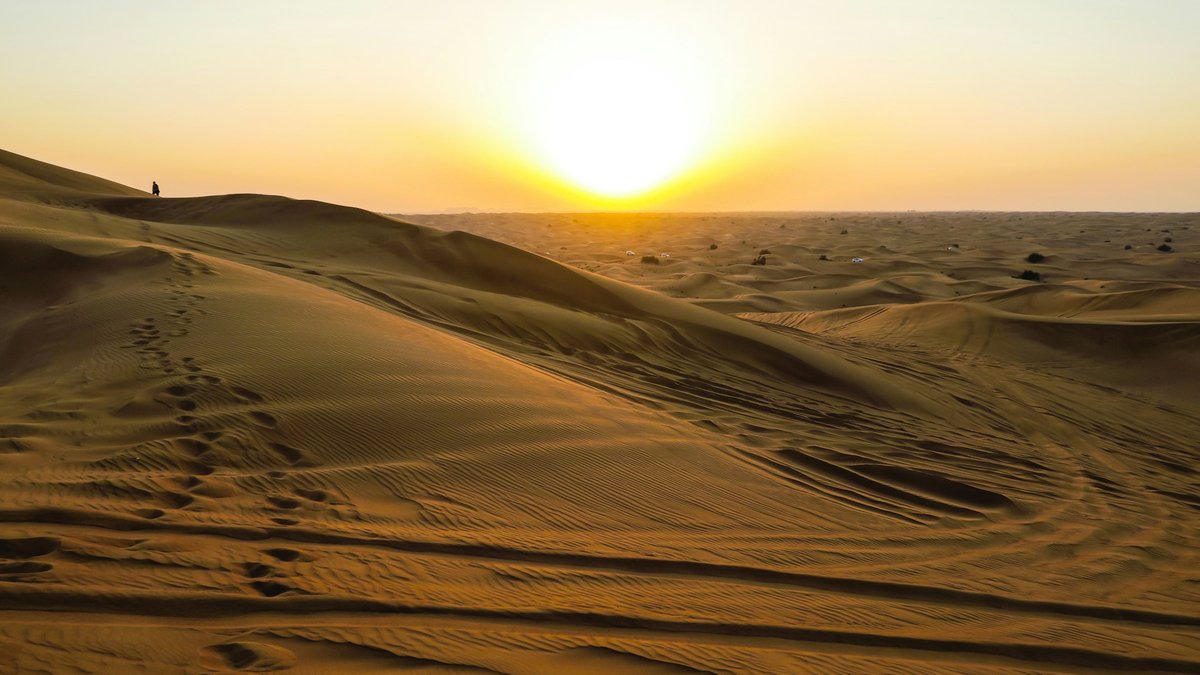 A molten sun hovers over rippled golden dunes in the Dubai desert, human footprints curving down the slip-face toward the foreground while a tiny silhouette stands on the distant crest