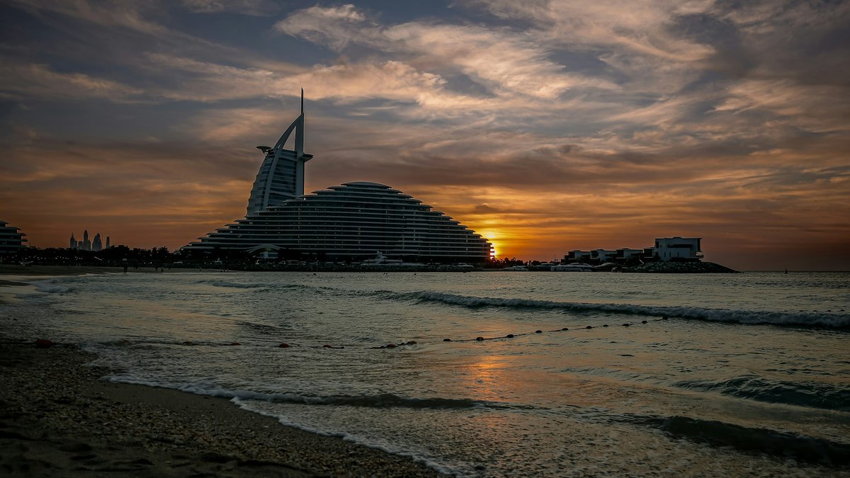 The sail-shape of Burj Al Arab rises beside the wave-curve of Jumeirah Beach Hotel as a brooding mackerel-sky sunset burns orange behind them, gentle waves lapping the empty Jumeirah Beach sand in the foreground