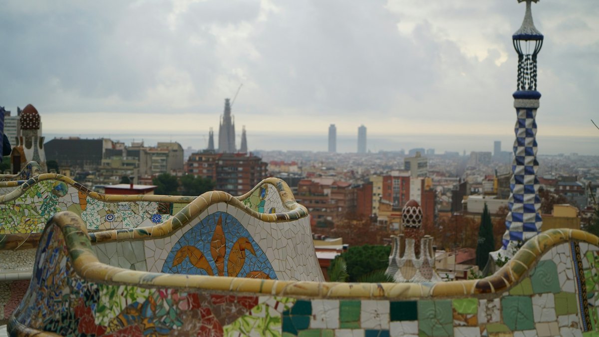 The serpentine trencadís bench at Park Güell, its hand-broken ceramic tiles in cobalt, ochre, and ivory snaking across a terrace that opens onto the Barcelona skyline