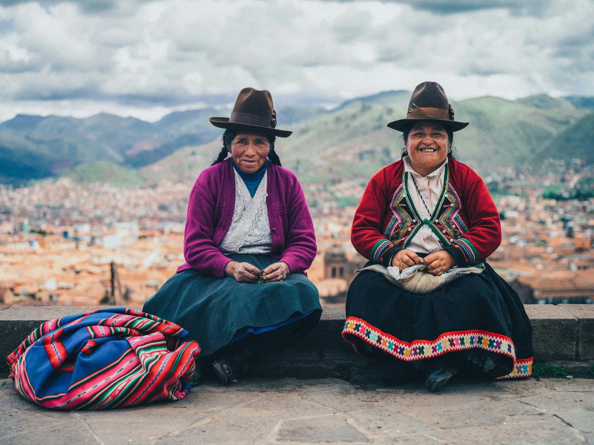 Two women in traditional hats sit overlooking a mountain city.