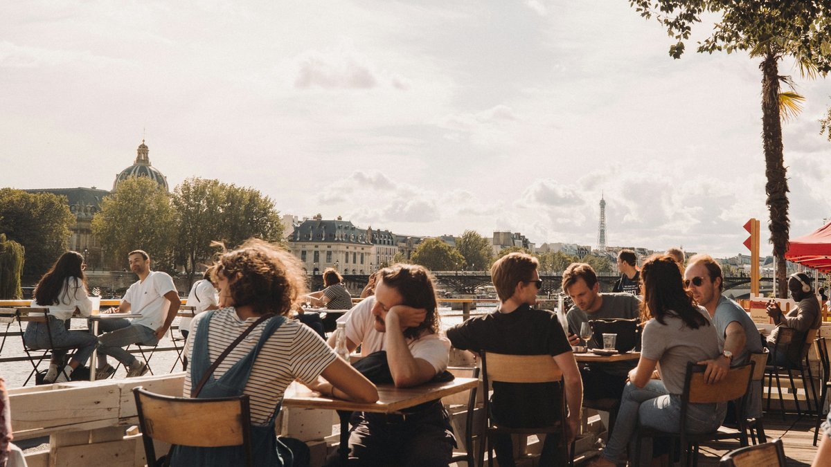 A Seine-side café in the afternoon, wooden chairs full of Parisians in slow conversation while the Eiffel Tower hazes into the distance