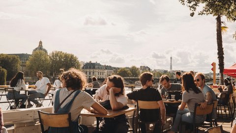 A Seine-side café in the afternoon, wooden chairs full of Parisians in slow conversation while the Eiffel Tower hazes into the distance