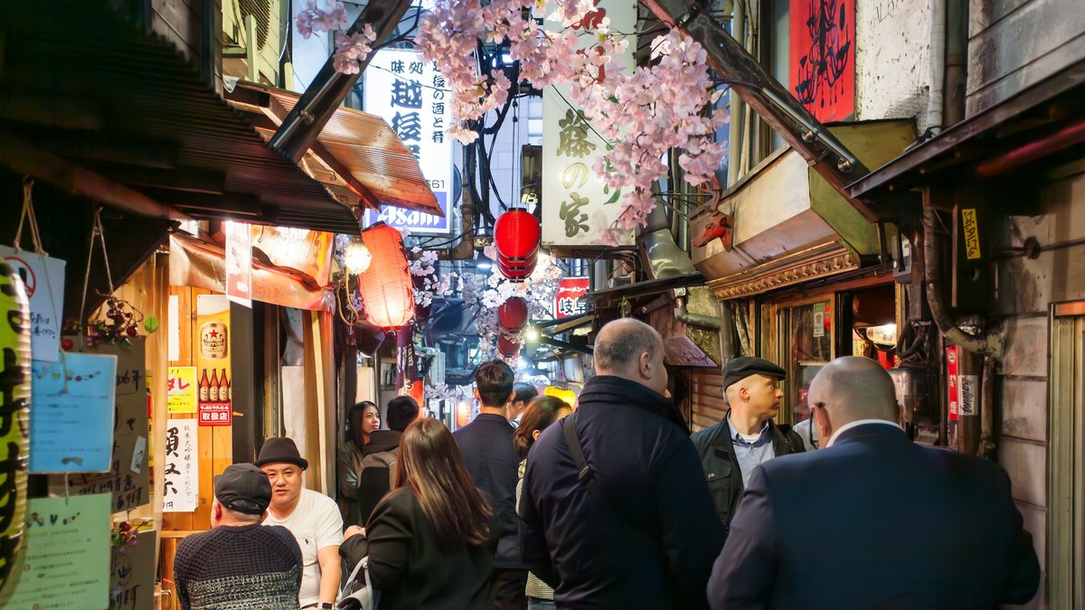 Salarymen drift between shoulder-width izakaya stalls in Omoide Yokocho, red paper lanterns and hand-painted signs drenching the narrow alley in a warm amber haze