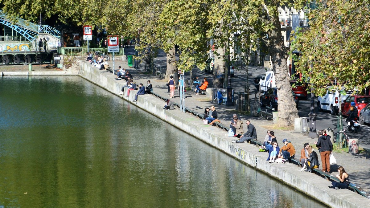 Parisians lounging along the stone banks of Canal Saint-Martin on a bright autumn afternoon, plane trees reflecting in the still green water