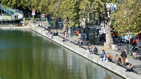 Parisians lounging along the stone banks of Canal Saint-Martin on a bright autumn afternoon, plane trees reflecting in the still green water
