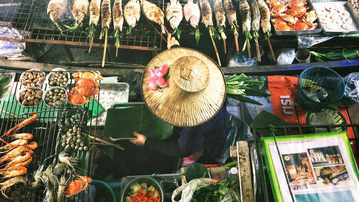 Overhead view of a Bangkok floating market stall, a vendor in a conical straw hat reaching for grilled fish, crab, and herbs arranged in vivid trays