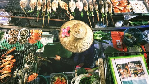 Overhead view of a Bangkok floating market stall, a vendor in a conical straw hat reaching for grilled fish, crab, and herbs arranged in vivid trays