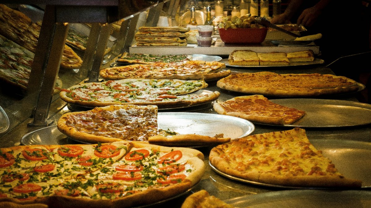 A New York slice-shop counter glowing under warm heat lamps, lined with dinner-plate pizzas — margherita with basil, plain cheese, pepperoni and white pies — while the pizzaiolo's hand reaches in to portion a slice