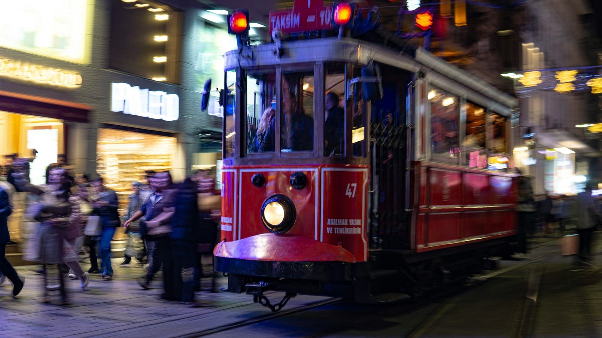 The vintage red İstiklal tram gliding down Taksim at twilight, illuminated shopfronts washing the surrounding crowd in warm violet and amber light