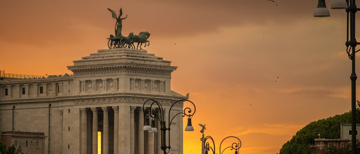 A golden sunset bathes Rome's terracotta rooftops and baroque domes, the Tiber's bends glimmering as the Eternal City fades into a warm, hazy horizon