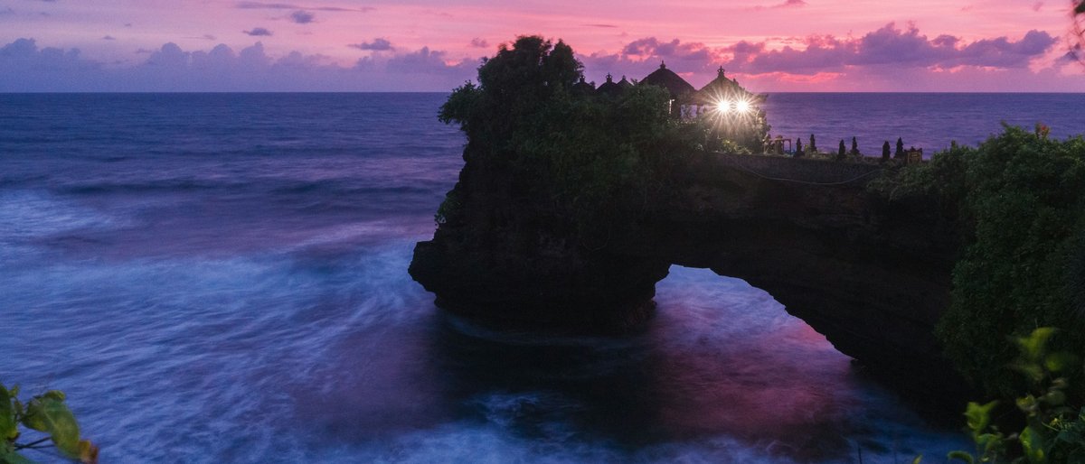 A small sea temple perched on a natural rock arch at Batu Bolong near Tanah Lot, silhouetted against a pink-and-violet twilight sky as long-exposure surf smooths the Indian Ocean into silk