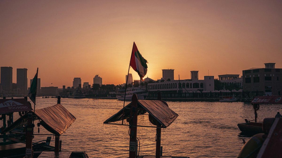 Wooden-canopy abra water-taxis moored along Dubai Creek at amber sunset, a UAE flag flying between the masts as the Bur Dubai skyline and Al Seef arches glow across the water
