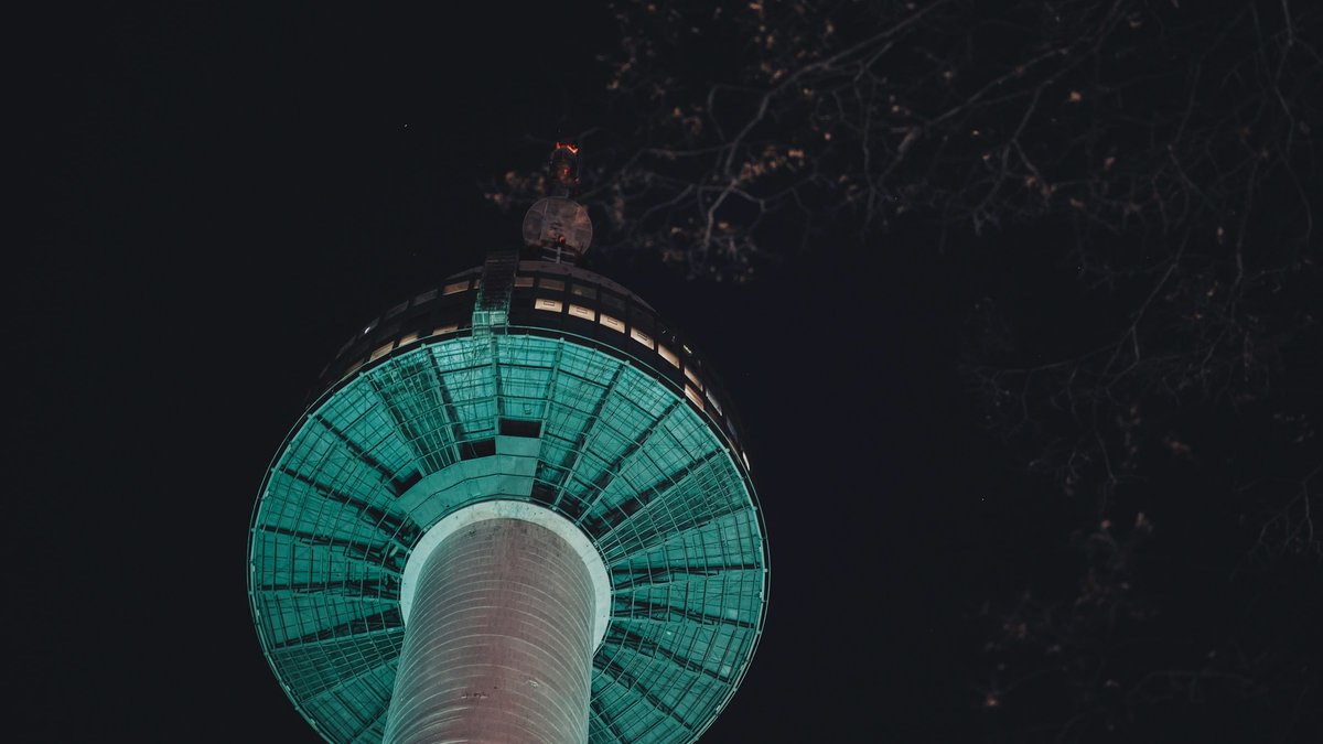A dramatic up-angle of N Seoul Tower's teal-lit observation deck rising from Namsan's dark forest canopy, bare cherry-blossom branches lacing the night sky around the spire