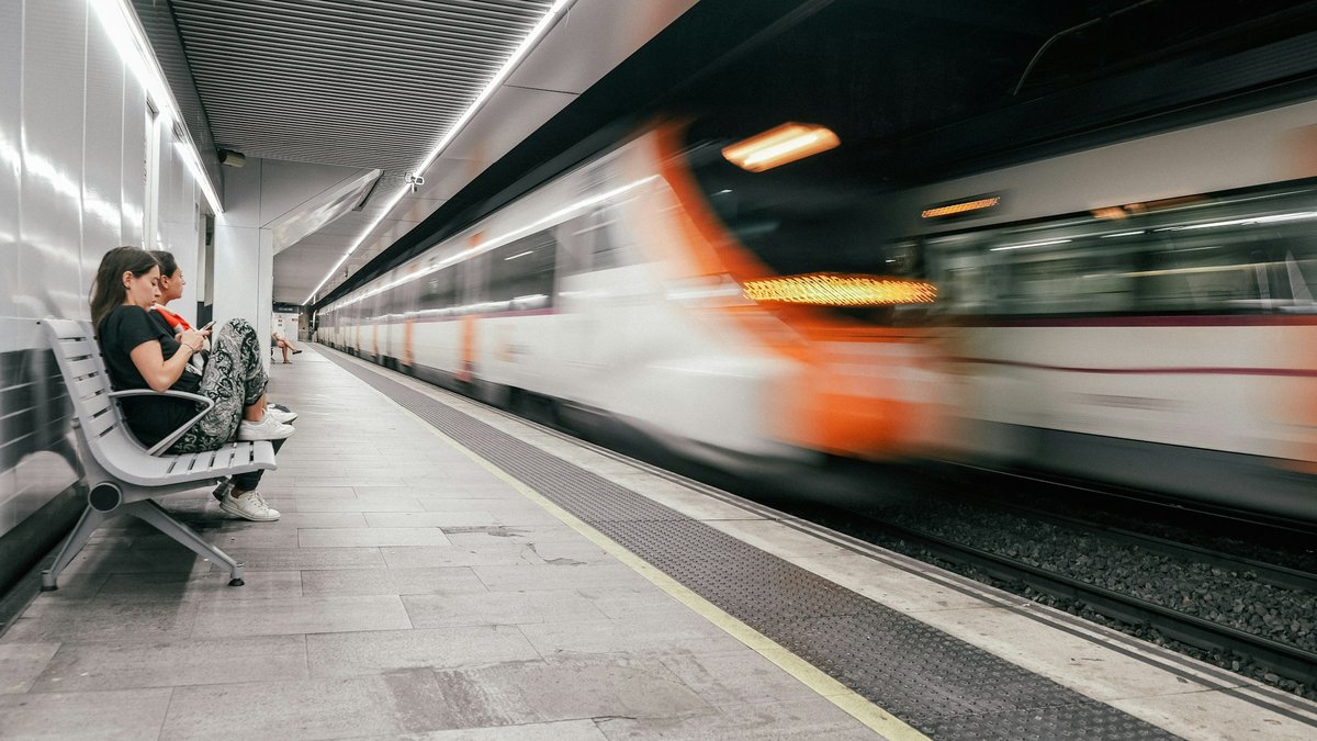 A Barcelona Rodalies commuter train streaks past an underground platform in a blur of white and orange light, a pair of travellers waiting calmly on the bench alongside