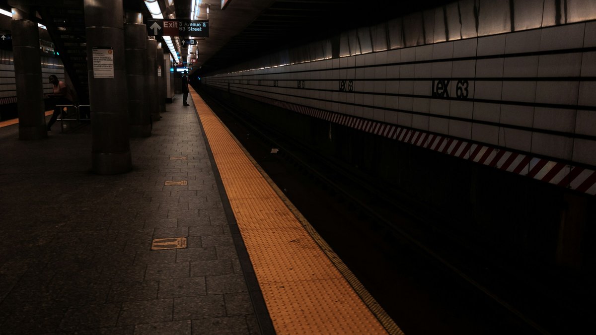 A moody MTA subway platform at the 63rd Street station, the yellow safety stripe and tile wall vanishing toward a distant Exit sign, lone figures small in the warm overhead light against a dark trackbed