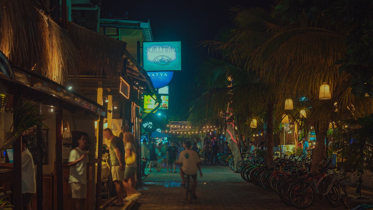 A Canggu-style gang at night — thatched warungs and neon bar signs glow teal and gold, rattan lanterns strung between palms, silhouetted travelers drifting between parked bicycles and scooters
