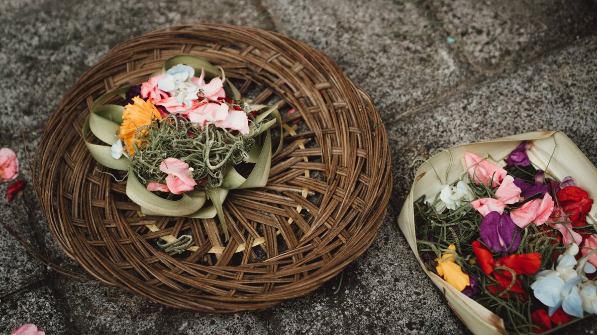 Two canang sari offerings rest on weathered temple stone — a woven rattan tray and a folded palm-leaf box filled with marigold, frangipani and pandan, a morning ritual for Balinese Hindus
