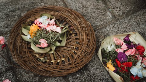 Two canang sari offerings rest on weathered temple stone — a woven rattan tray and a folded palm-leaf box filled with marigold, frangipani and pandan, a morning ritual for Balinese Hindus