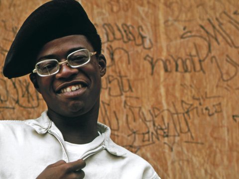 a man wearing glasses and a hat standing in front of a wooden wall