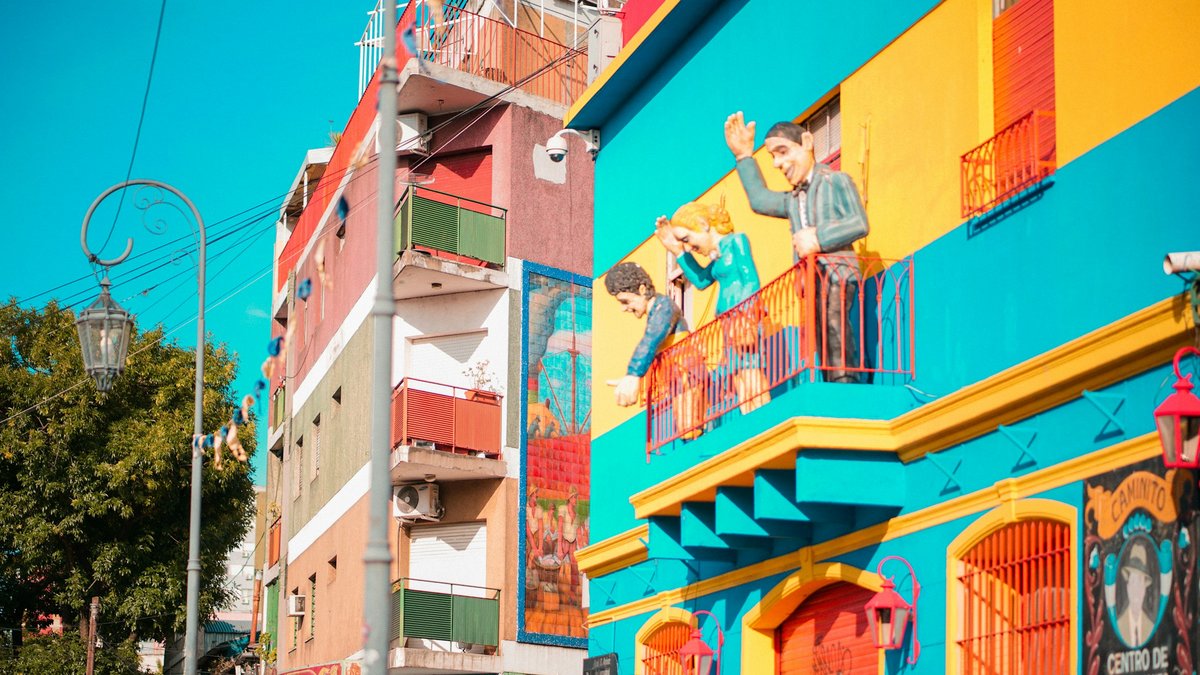A Caminito corner in La Boca painted yellow, red and turquoise, its balcony crowded with caricature figures of Maradona, Evita and Carlos Gardel, a wrought-iron streetlamp glowing beside the Centro de Exposiciones sign